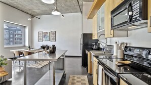 Une cuisine moderne équipée d’appareils électroménagers en acier inoxydable, un coin repas avec une longue table et une vue sur la ville grâce à de grandes fenêtres.