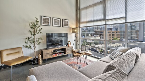 Un salon moderne avec une grande fenêtre, un canapé, un téléviseur et une petite table d’appoint en bois.