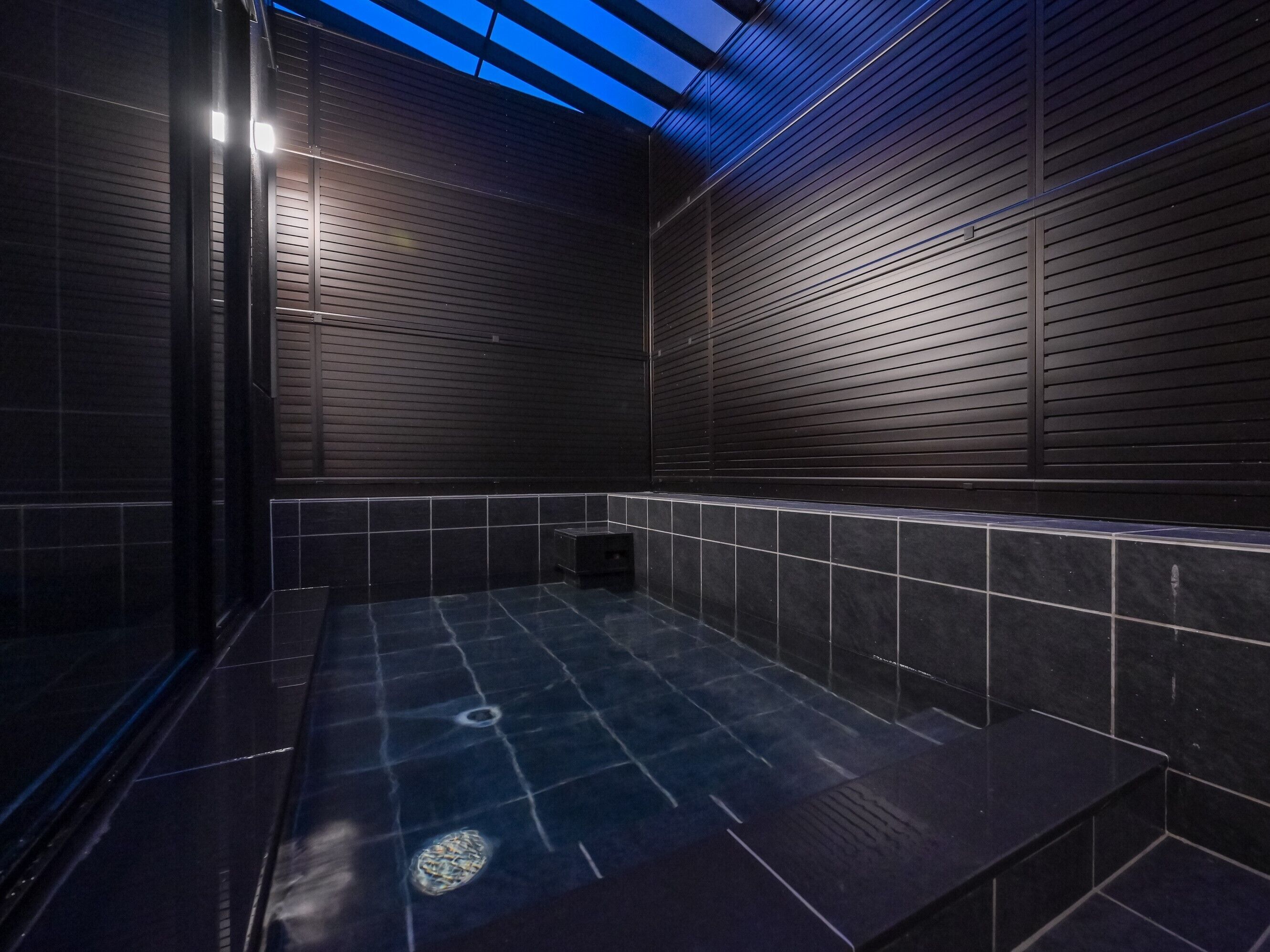 Une salle de bain moderne avec des murs recouverts de carreaux foncés, une douche en verre et un puits de lumière.