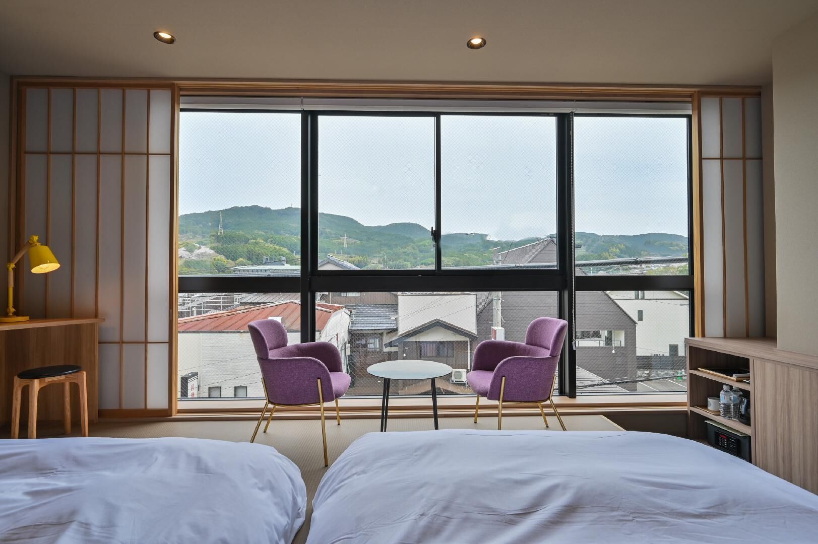 A bedroom with two beds, a small table, two purple chairs, a yellow lamp, and a large window with a view of buildings and hills.