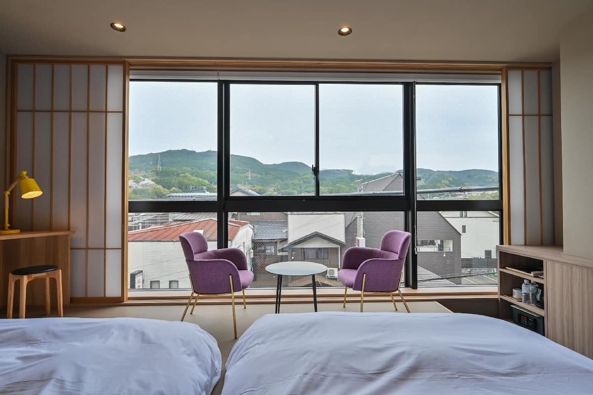 A bedroom with two beds, a small table, two purple chairs, a yellow lamp, and a large window with a view of buildings and hills.