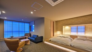 A modern hotel room with a large bed, a seating area with a sofa and armchair, a small table, and a large window with a view.