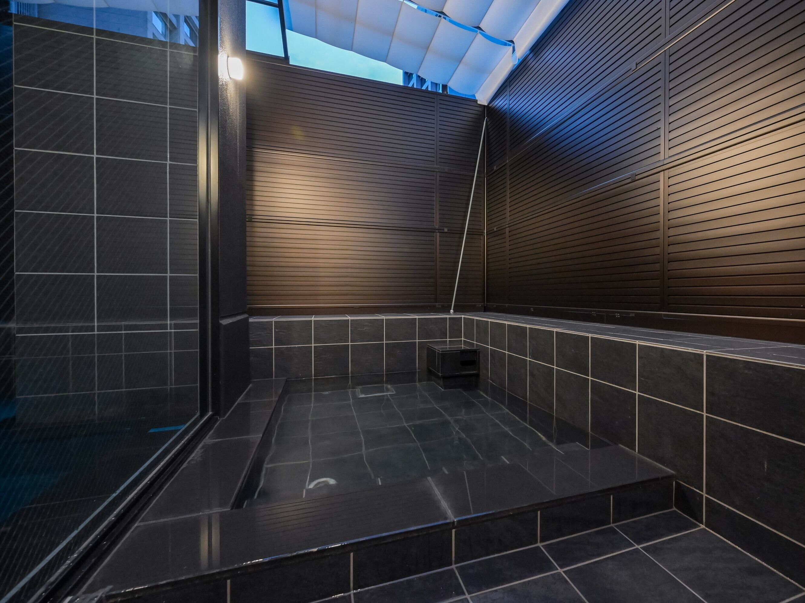 A modern shower area with dark tiles, a wooden slatted wall, and a skylight.