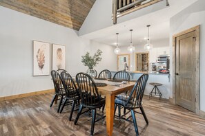 A dining area with a wooden table and chairs, a vase with greenery, and a kitchen in the background.