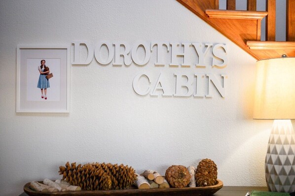 Un letrero que dice 'DOROTHY'S CABIN' con un marco que contiene una foto de una mujer y un arreglo decorativo de piñas sobre una mesa.