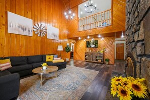 A spacious living area with a wooden floor, a black sofa, a round coffee table, and a large rug. There is a staircase leading to an upper level with a balcony.