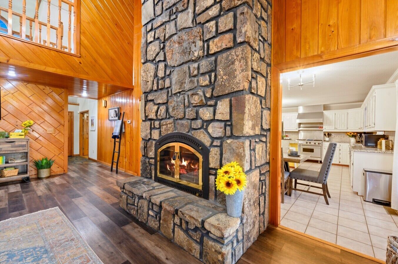 A stone fireplace with a fire, wooden floors, and a kitchen area with white cabinets.