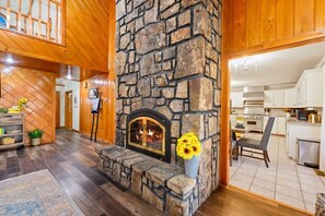 A stone fireplace with a fire, wooden floors, and a kitchen area with white cabinets.