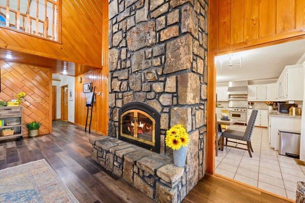 A stone fireplace with a fire, wooden floors, and a kitchen area with white cabinets.
