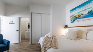 Una camera d'hotel con un letto, una poltrona blu, un quadro appeso al muro e una porta con la scritta '20B Beachcomber'.