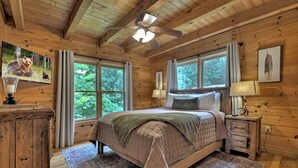 Quarto de cabana de madeira com cama, duas janelas, ventilador de teto e televisão na parede.