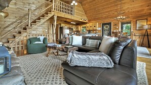 Interior de cabana de madeira com escada, sala de estar com sofá de couro, área de refeições e cozinha.