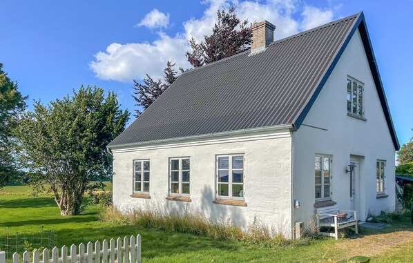 Une maison blanche à deux étages, avec un toit gris, une clôture en bois et un banc à l’extérieur.