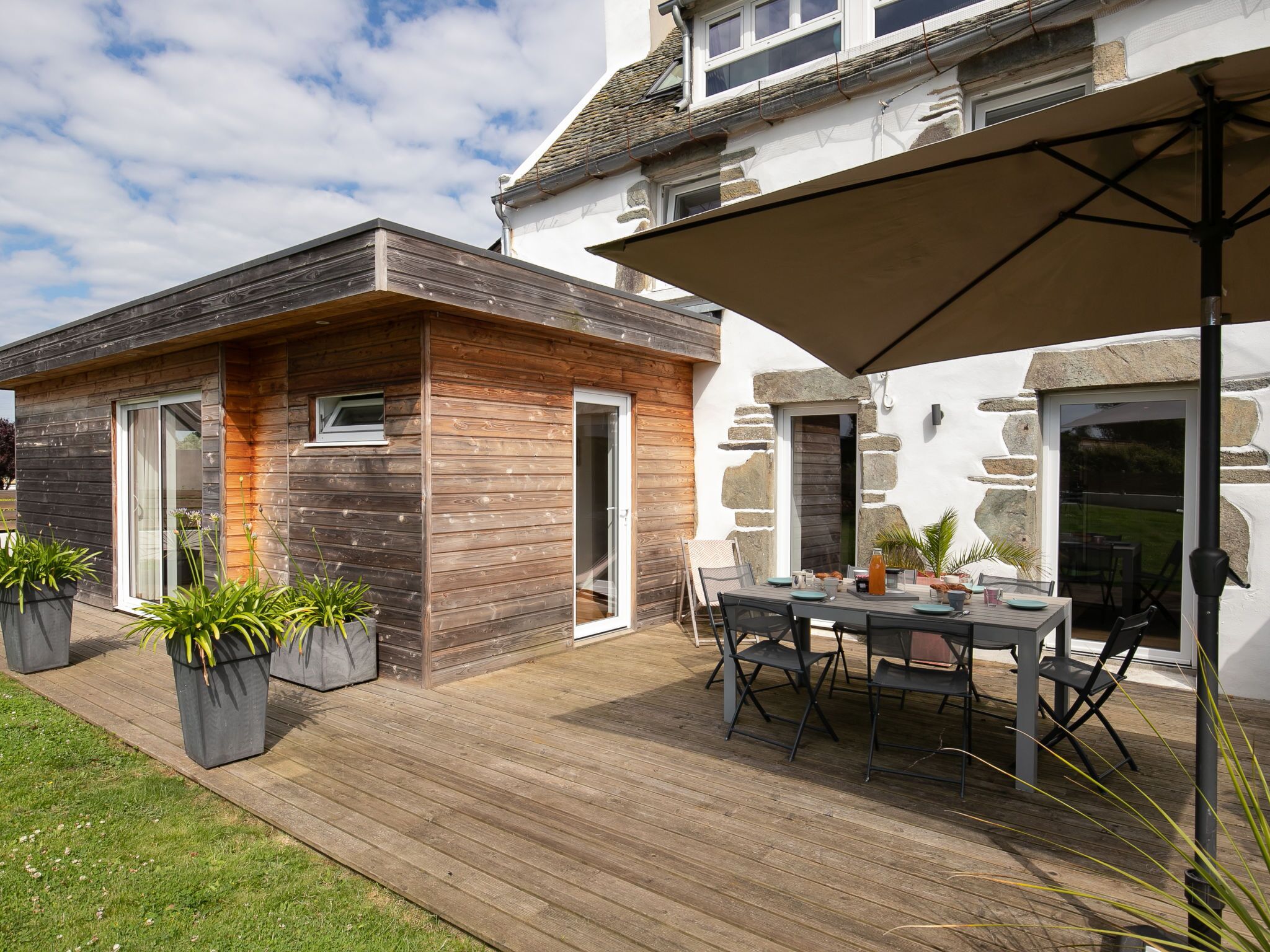 Une maison moderne dotée d’une terrasse en bois, d’un coin repas avec mobilier d’extérieur et d’un mur en pierre.