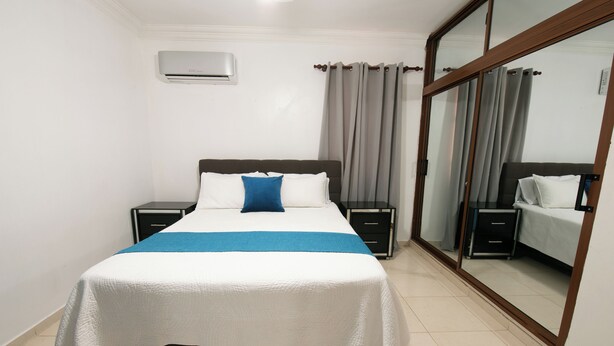 A neatly made bed with a white and blue bedspread, two bedside tables, a nightstand, a large mirror, and a sliding glass door. - Próximo a unión medica 2 hab. (Santiago de los Caballeros)