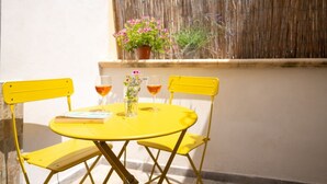 A yellow table with two chairs, a potted plant, and two glasses of red wine on a sunny patio.