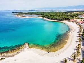 An aerial view of a coastal area with a beach, clear turquoise waters, and a backdrop of mountains.