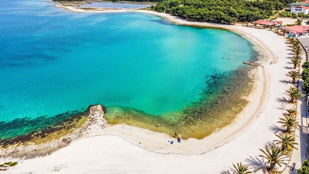 Ein Luftbild einer Küstenlandschaft mit Strand, klarem türkisblauem Wasser und einer Bergkulisse im Hintergrund.