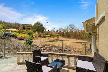 A balcony with wicker furniture and a view of a landscaped area with trees and a building in the background.