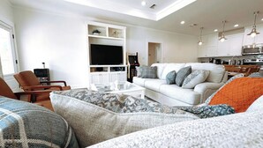 A modern living room with a white sofa, a glass coffee table, and patterned cushions. There is a TV on a white cabinet, and a kitchen area with white cabinets and stainless steel appliances. - Elegant home 180 steps from the Beach, 2 King beds, Arcade (Bolivar Peninsula)