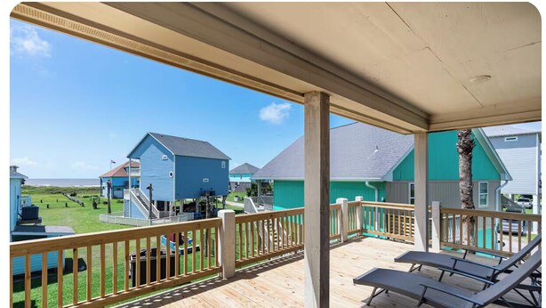 A covered outdoor deck with a wooden railing, a pool, and a view of a blue house and a grassy area. - Elegant home 180 steps from the Beach, 2 King beds, Arcade (Bolivar Peninsula)
