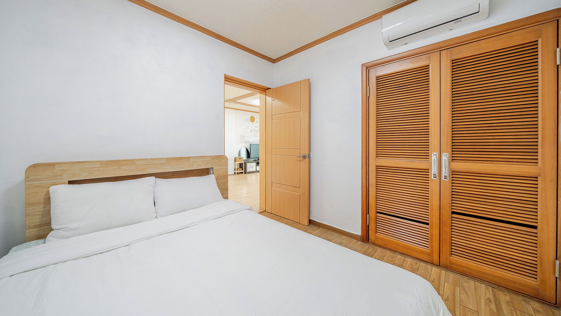 Une chambre avec une tête de lit en bois, un lit avec une literie blanche, une armoire en bois dotée de portes à persiennes et un climatiseur fixé au mur.
