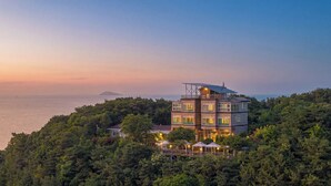 A multi-story building with a rooftop terrace, surrounded by dense forest, overlooking a body of water.