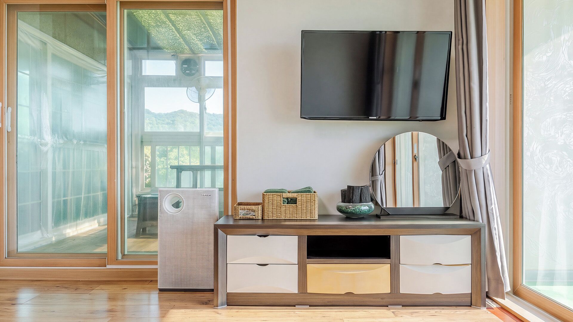 A room with a wooden floor, a TV mounted on the wall, a large window with curtains, and a sliding glass door leading to an outdoor area.