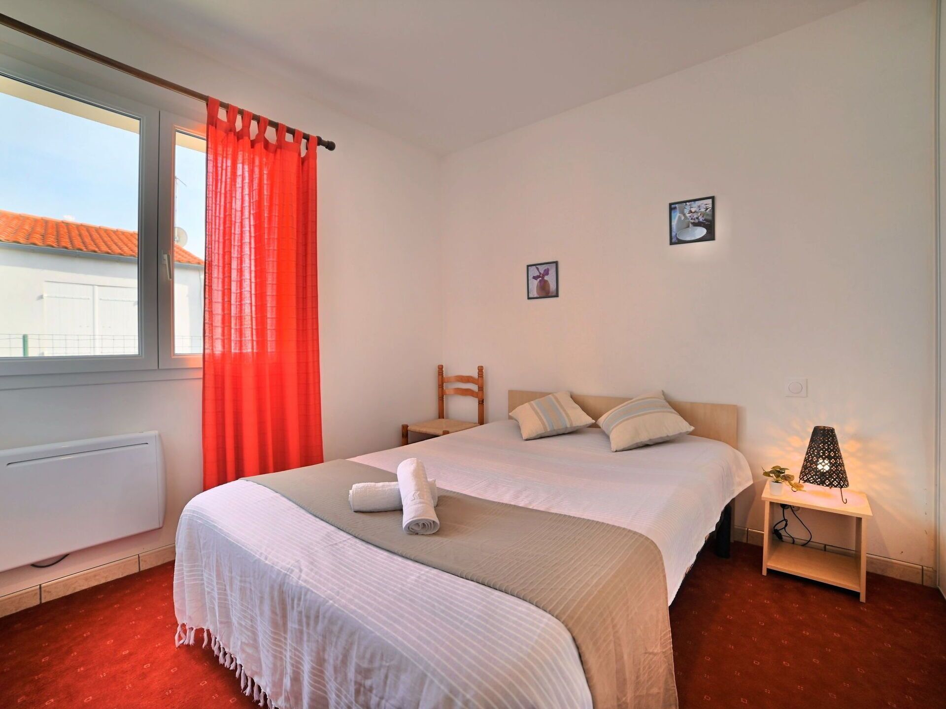 A hotel room with two beds, a window with red curtains, a chair, a nightstand with a lamp, and a small picture on the wall.