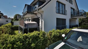 Um edifício residencial moderno de dois andares com área externa coberta, um carro estacionado na frente e um céu azul claro.