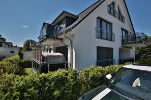 Um edifício residencial moderno de dois andares com área externa coberta, um carro estacionado na frente e um céu azul claro.