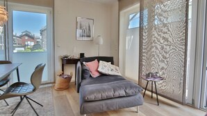 Quarto moderno com uma cama, uma escrivaninha e uma cadeira. Há uma janela com vista para prédios.
