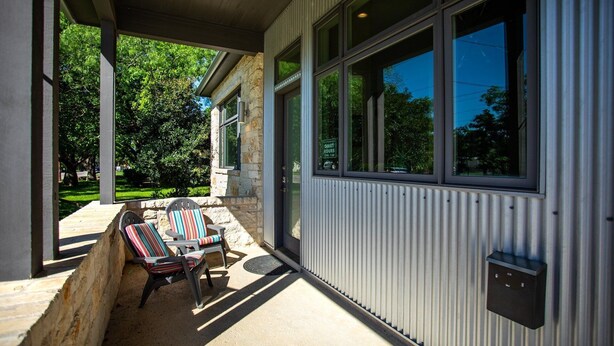 A covered patio area with two striped chairs and a table, large windows, and a stone wall. - Sta-Haus | Wonderful Location (Fredericksburg)