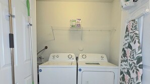 Une buanderie avec deux machines à laver à chargement frontal blanches, une étagère sur laquelle se trouve une boîte de feuilles d’adoucissant et un balai appuyé contre le mur.