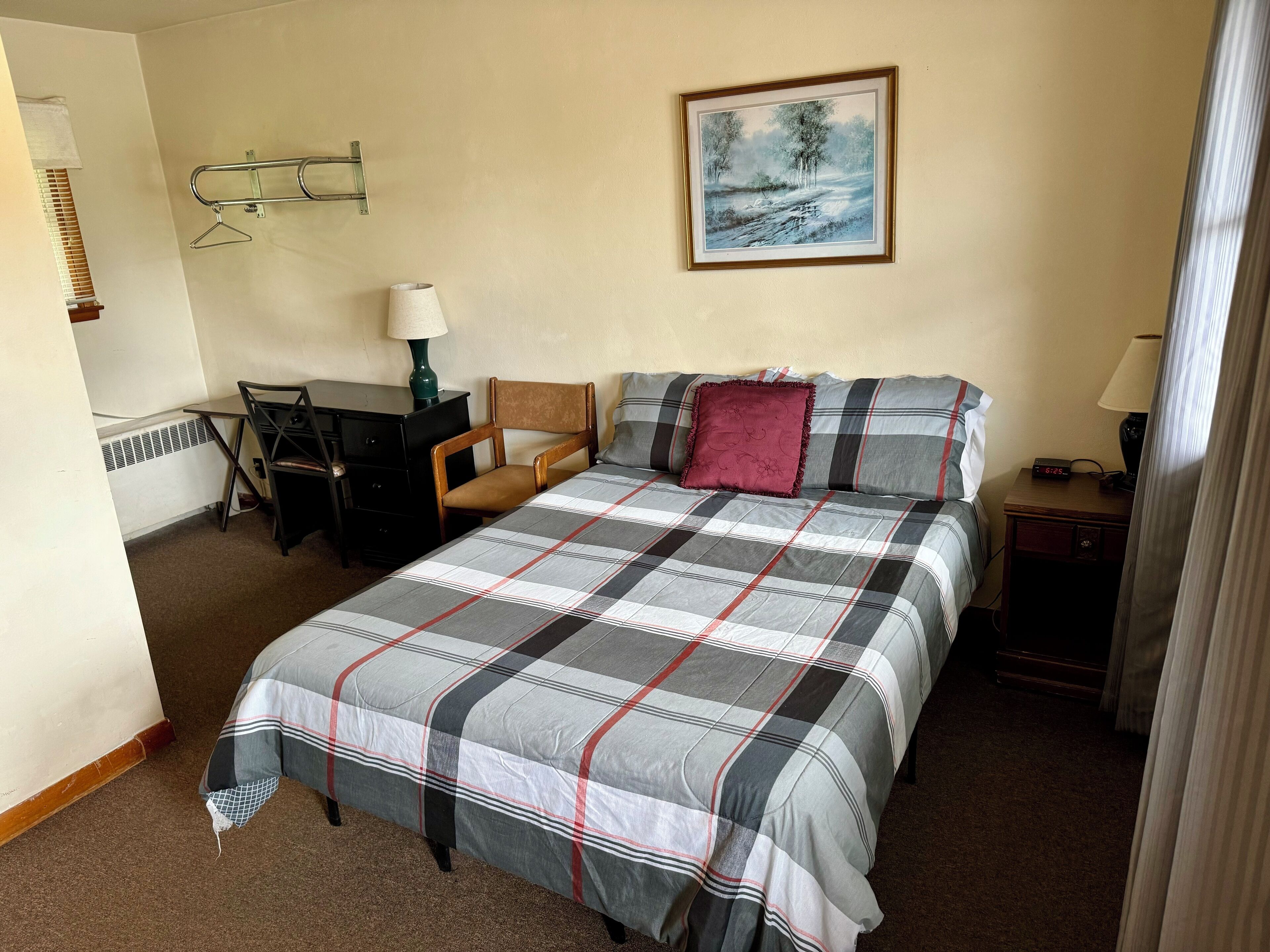 A hotel room with a bed, a desk, a chair, a painting on the wall, and a nightstand with a lamp.