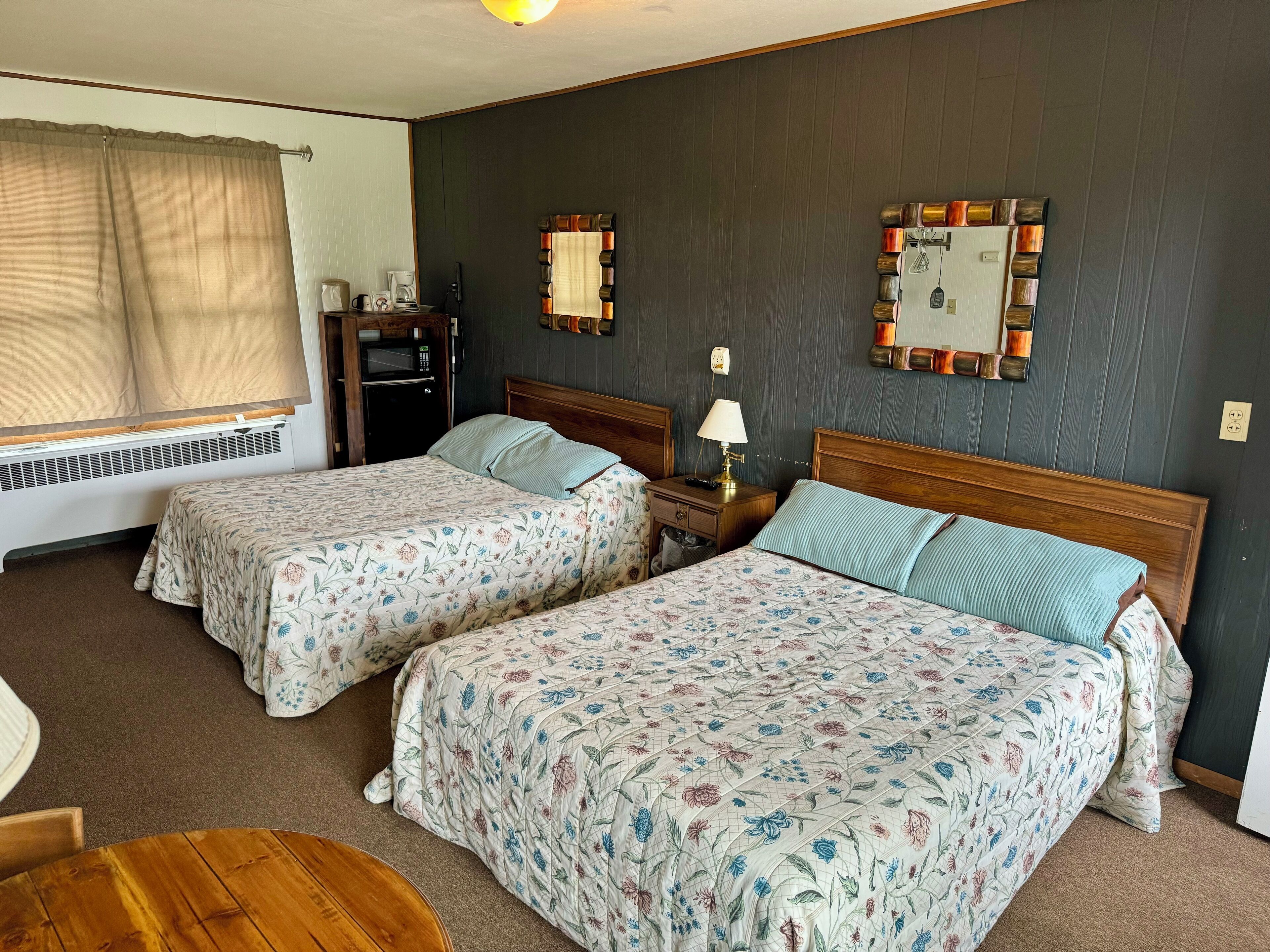 A hotel room with two beds, a wooden headboard, a bedside table with a lamp, a small table, and a window with curtains.