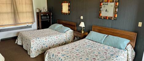 A hotel room with two beds, a wooden headboard, a bedside table with a lamp, a small table, and a window with curtains.