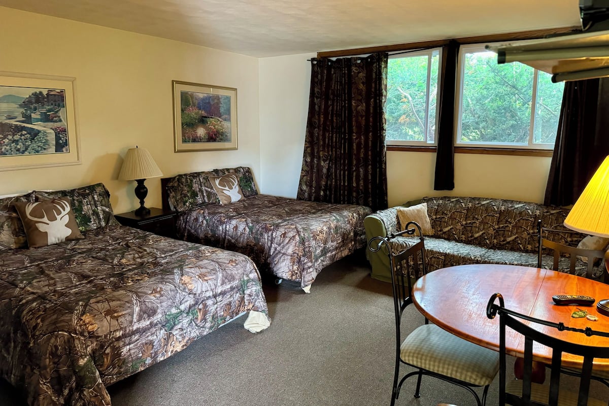 A room with two beds, a table, and chairs. There are two lamps, a window with curtains, and two framed pictures on the wall.