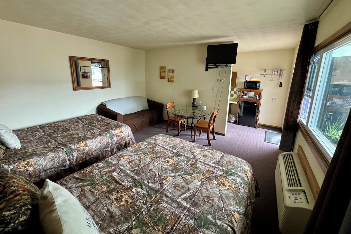A room with two beds, a small dining table with chairs, a TV mounted on the wall, and a kitchenette area.