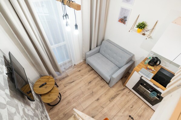 A modern living room with a grey sofa, wooden flooring, a small round table, and a kitchen area with a stove and coffee maker.