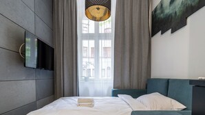 A modern hotel room with a bed, a TV mounted on the wall, a window with curtains, and a painting on the wall. - Żwirki i Wigury Apartment #2 (Katowice)