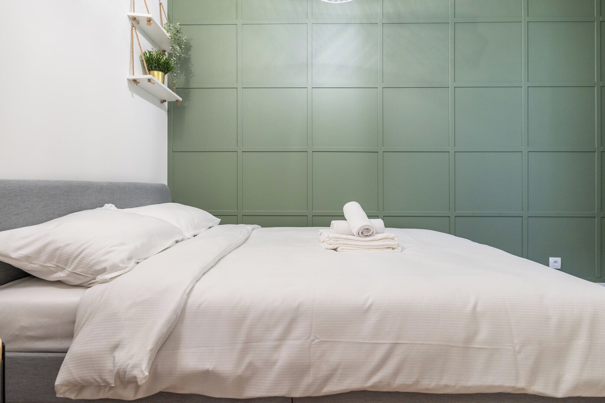 Ein ordentlich bezogenes Bett mit weißen Leinentüchern und einem zusammengerollten Handtuch, ein graues Sofa und eine grüne Wand mit Rastern.