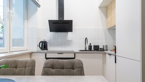 A modern kitchen with white cabinets, a black range hood, a kettle, a coffee maker, and a sink. There are two chairs and a table in front of the kitchen. - Żwirki i Wigury Apartment #3 (Katowice)