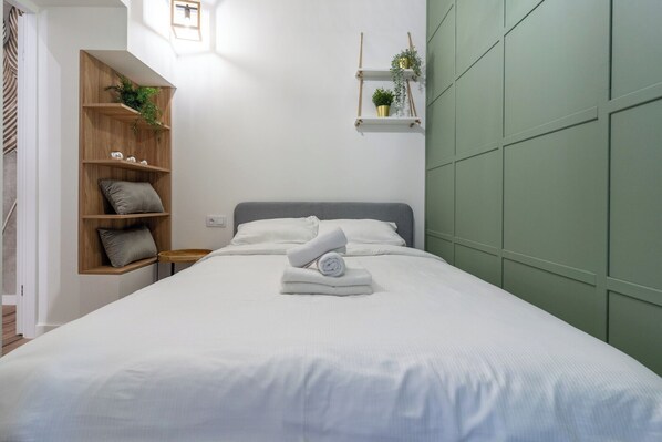 A neatly made bed with white linens and rolled towels, a wooden shelf unit with cushions, and a green padded wall.