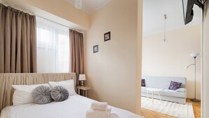 A neatly made bed with pillows, a bedside table with a lamp, a sofa, and a floor lamp in a well-lit room with a large window and curtains. - Rondo ONZ Standard Studio (Warszawa)