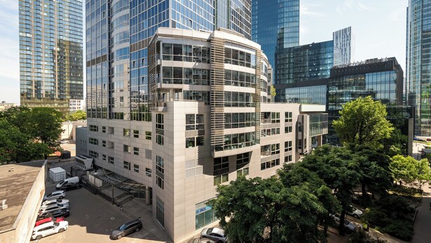 A modern building with a curved facade, large windows, and a parking lot with several cars. - Rondo ONZ Standard Studio (Warszawa)