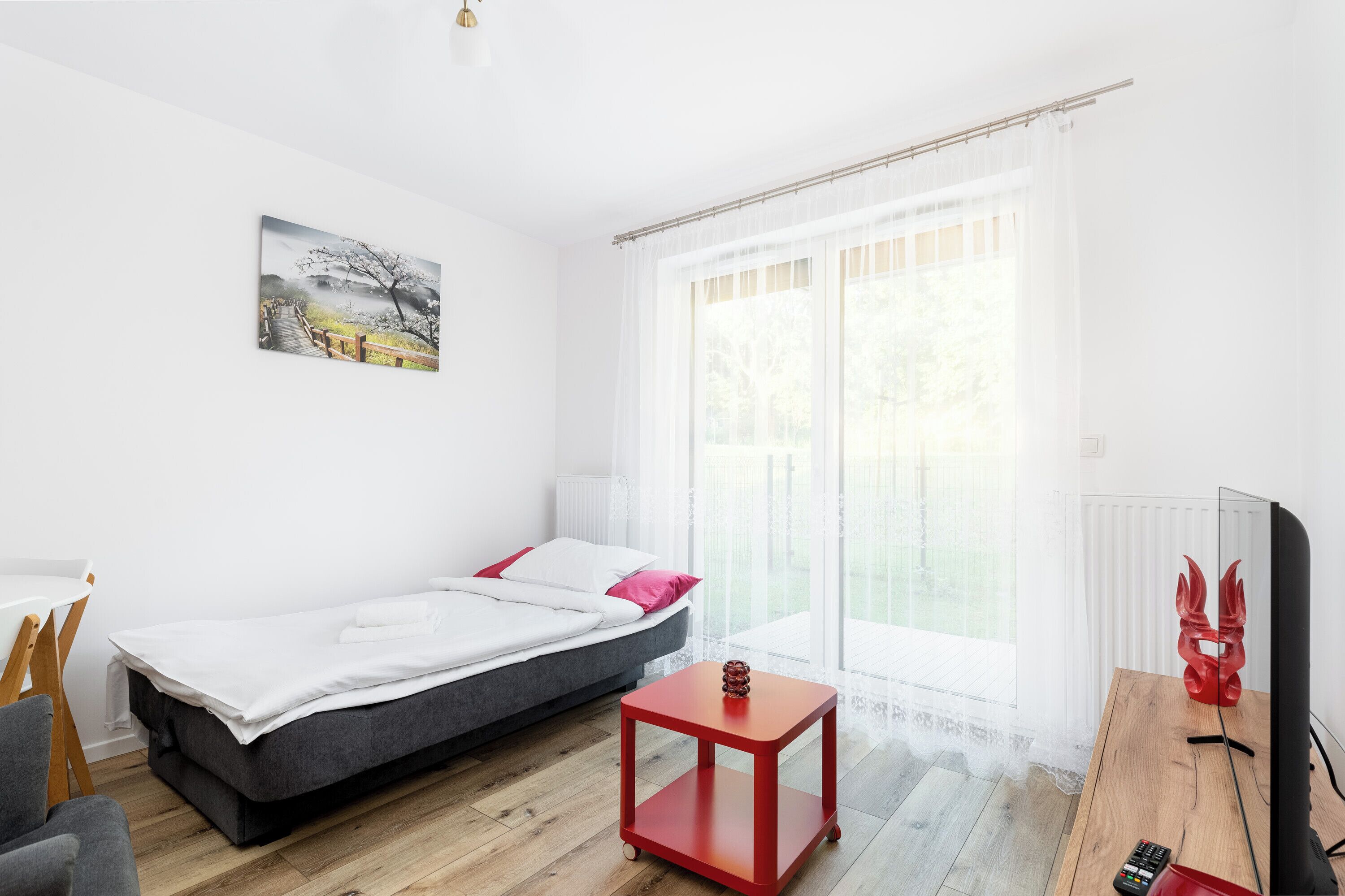 Um quarto moderno com uma cama, uma pequena mesa vermelha, uma televisão e uma janela com cortinas translúcidas.