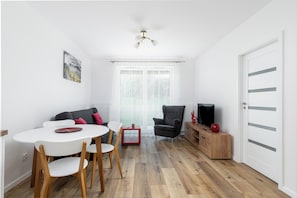 A modern living room with a white dining table, wooden chairs, a grey sofa, a black armchair, and a TV on a wooden cabinet.
