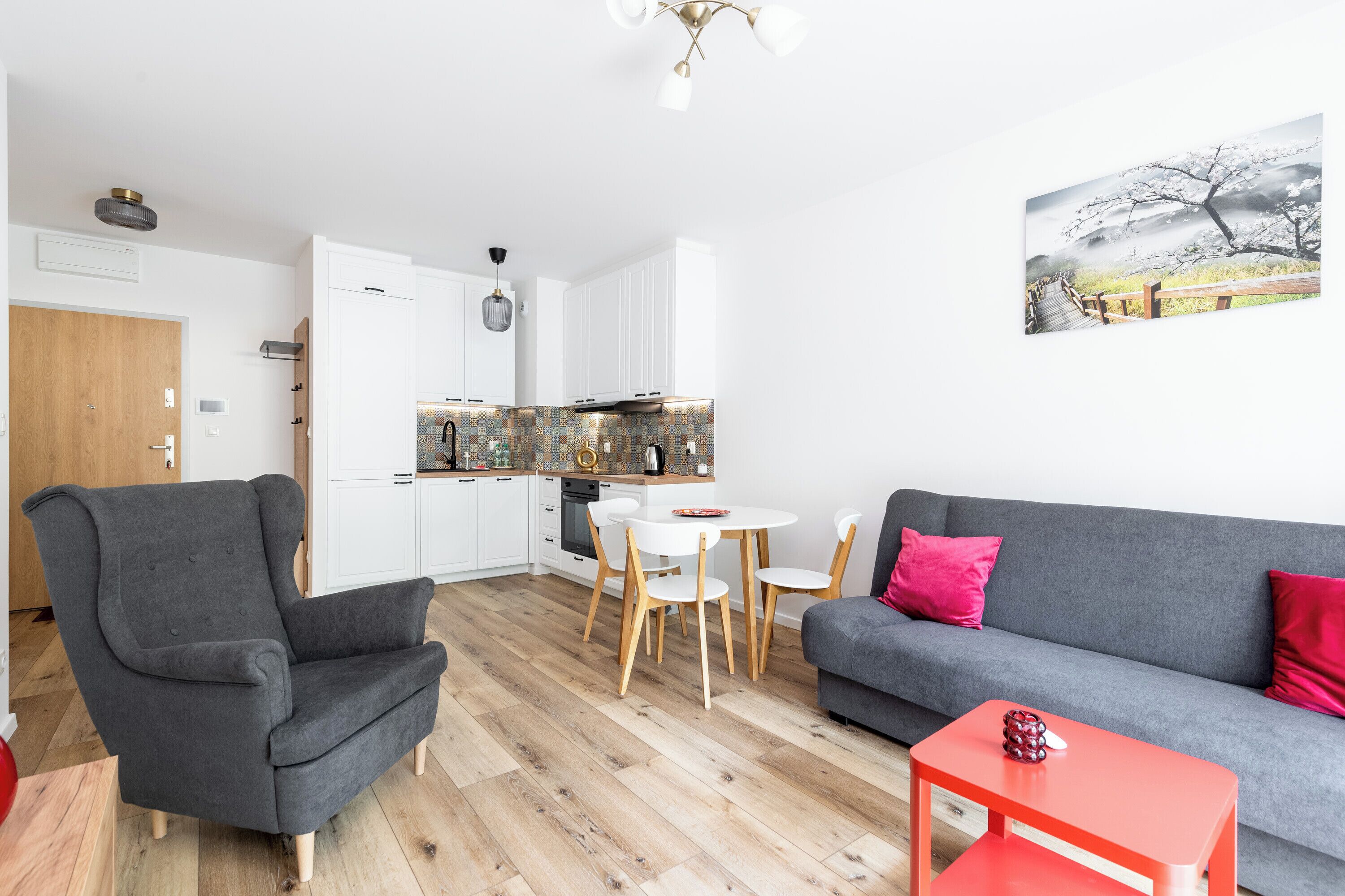 Een moderne woonkamer met een grijze bank, een rode bijzettafel en een grijze fauteuil. In de achtergrond is een keuken met witte kasten en een houten aanrecht te zien.
