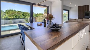 Een moderne keuken met een groot eiland, een eethoek en uitzicht op een zwembad en groen.